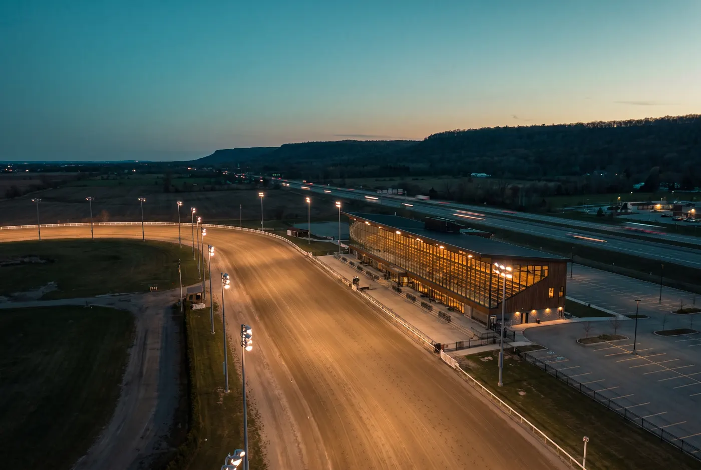 Illustration : vue de drone au crépuscule sur une piste de courses attelées vide, éclairée par des projecteurs, avec une longue tribune et une autoroute au loin